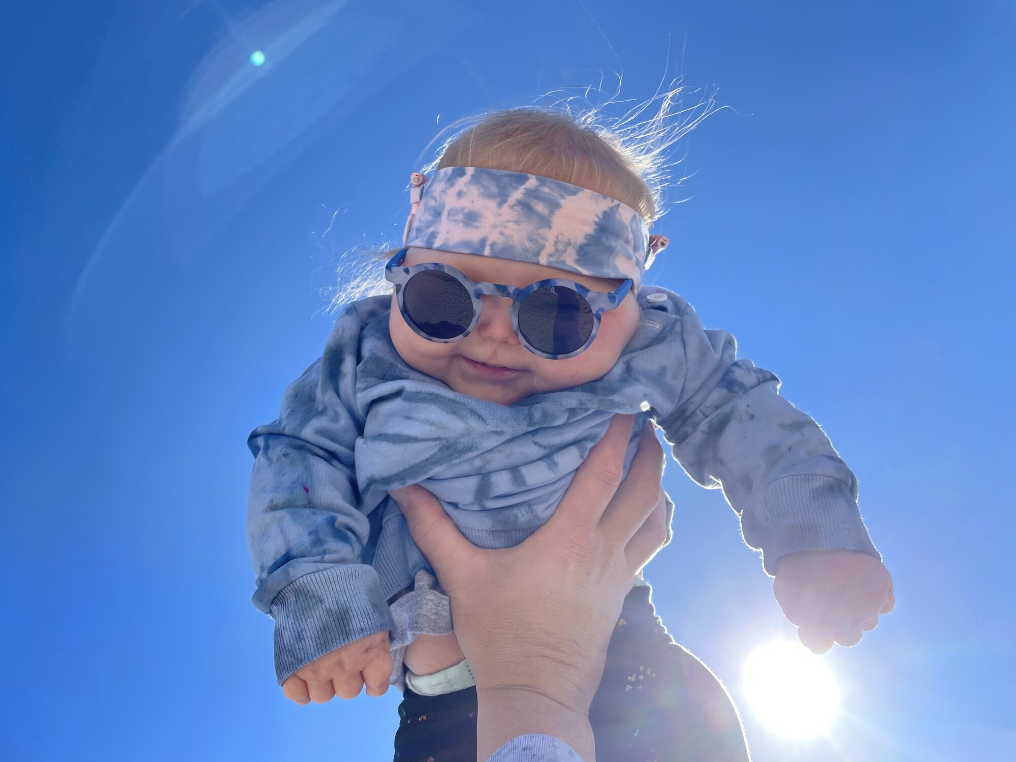 A baby, wearing sunglasses and a headband, being held in the air.
