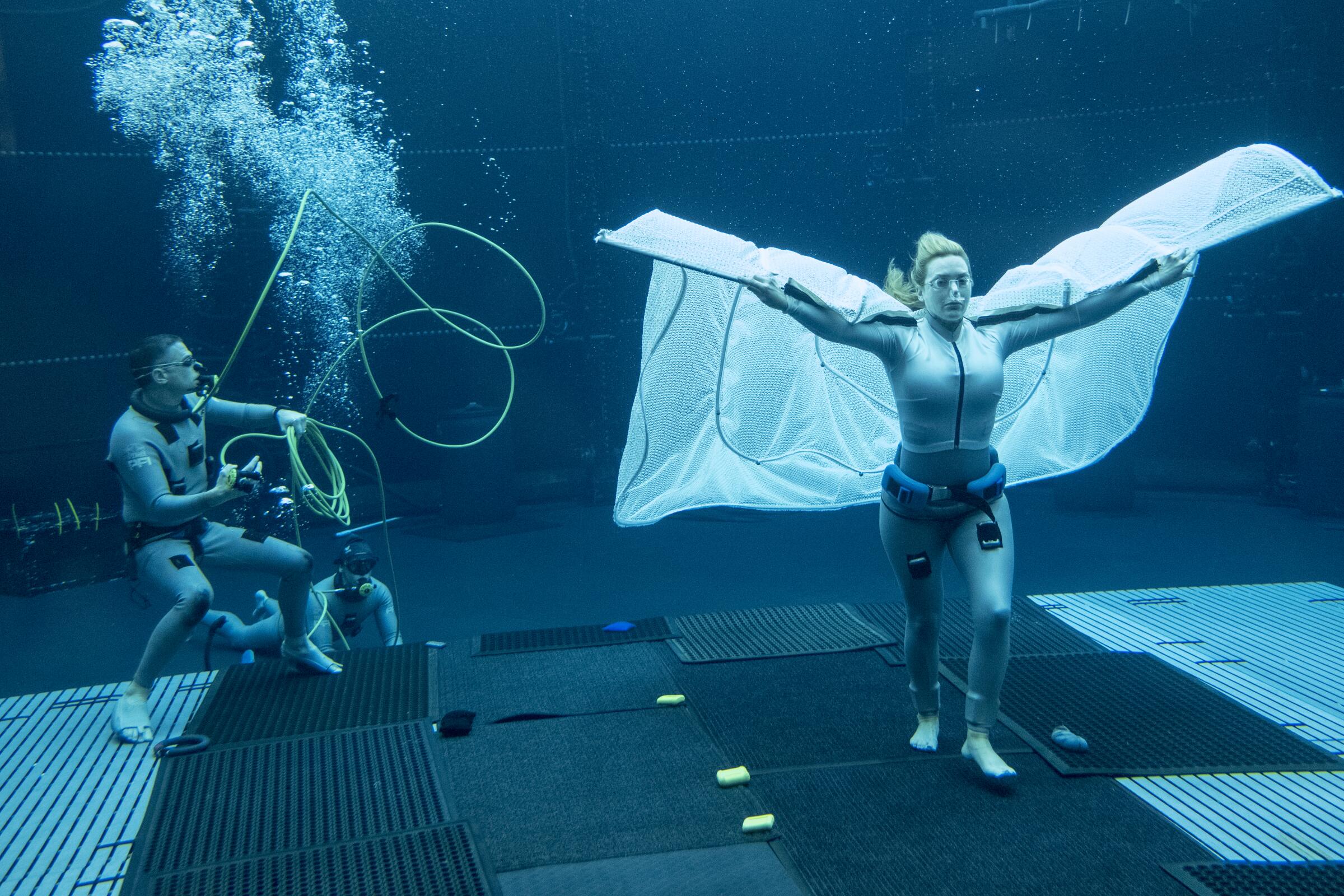 Kate Winslet is filmed walking underwater with a cape-like fabric stretched behind her.