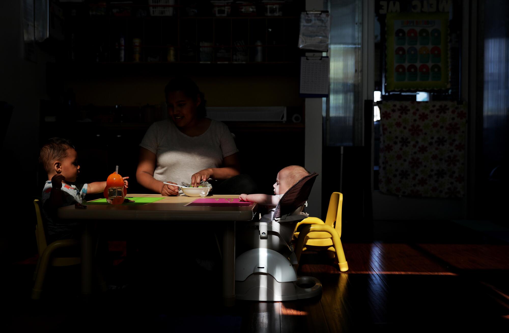 A baby seated at a children's table in a darkened room visible through a sliver of light.