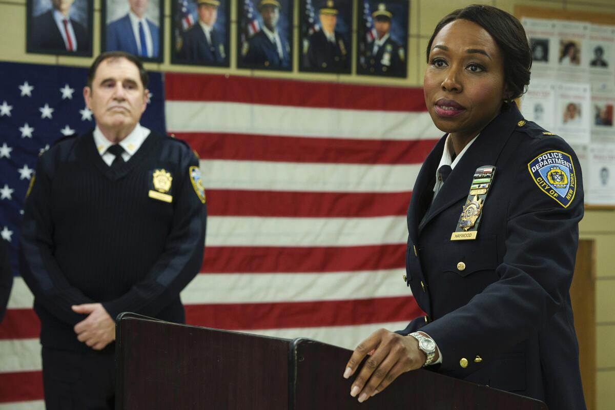A male police officer stands in front of an American flag, looking at a female police officer standing at a lectern.