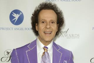 A man with short curly hair in a purple suit with a bedazzled trim and a matching tie smiling with his mouth open