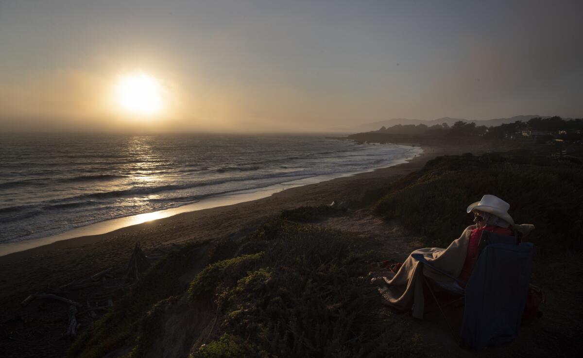Wearing a rimmed hat and wrapped in a blanket, a seated visitor watches the sun set at a beach.