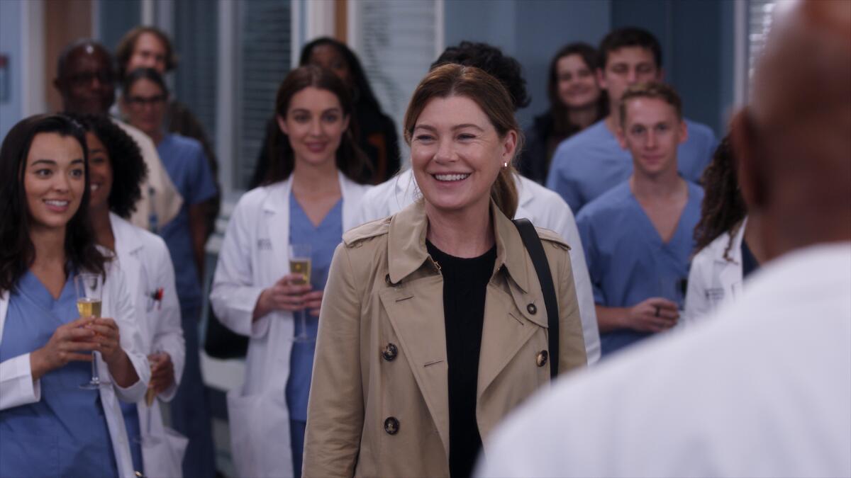 A woman in a brown trenchcoat is applauded by people in blue scrubs and white lab coats in a scene from "Grey's Anatomy."