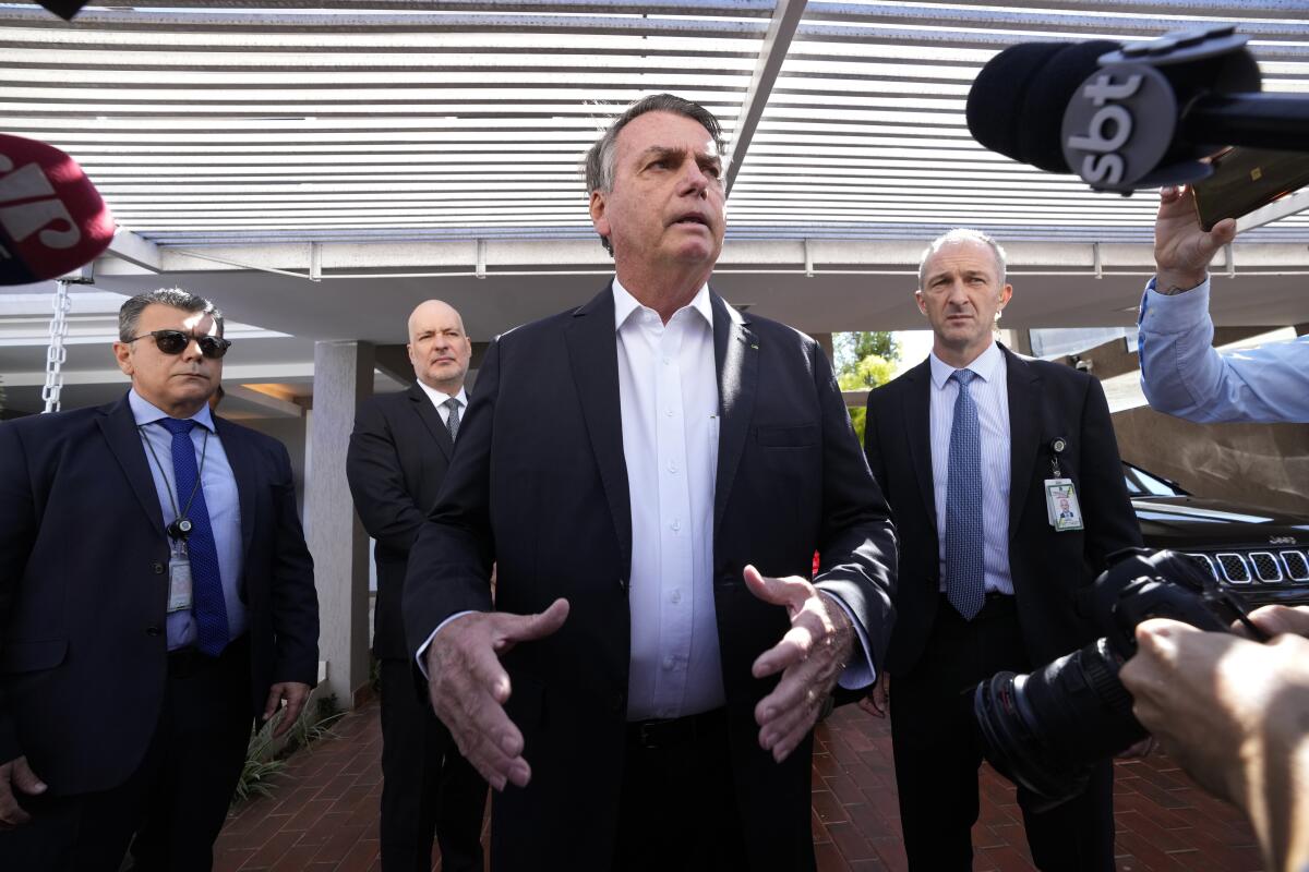 A man in a dark jacket and white shirt gestures with his hands. Behind him are three men in suit and tie