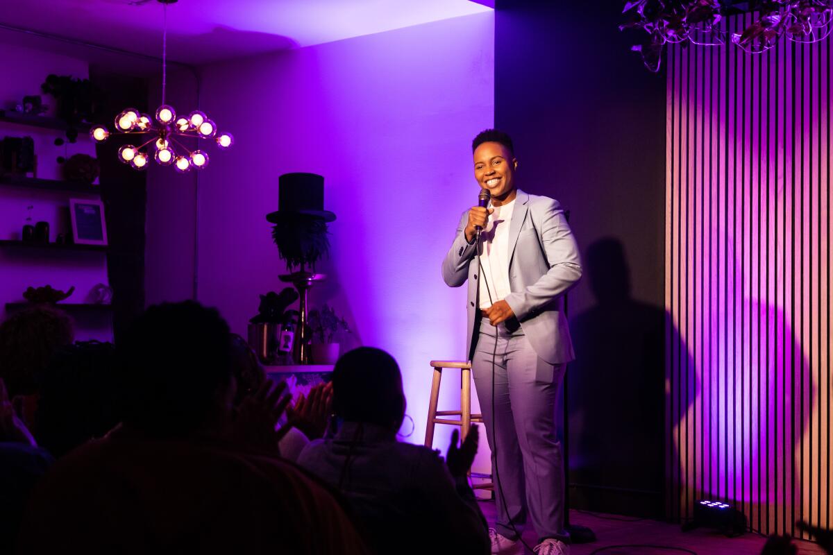 A woman in a gray suit holds a microphone on a stage in a room lit with purple lights