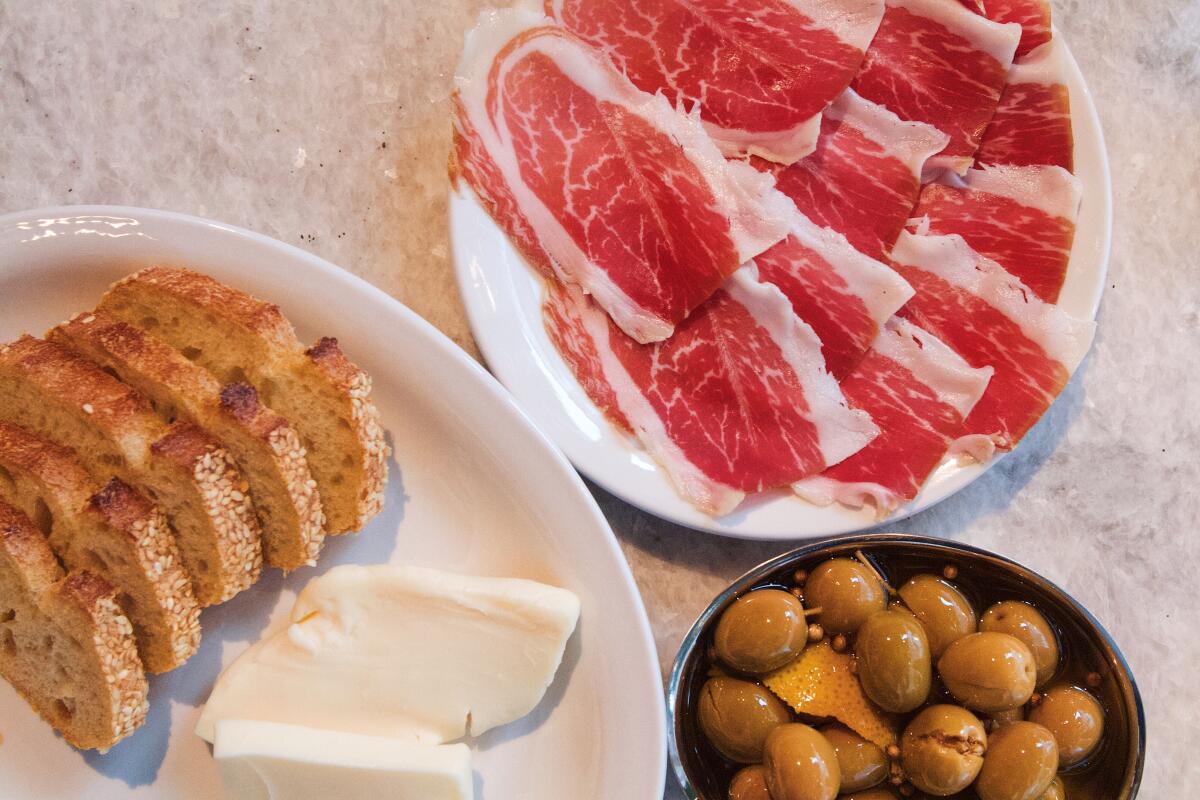 An overhead photo of slices of cured ham, a dish of green olives, and a plate with sliced baguette and cheese