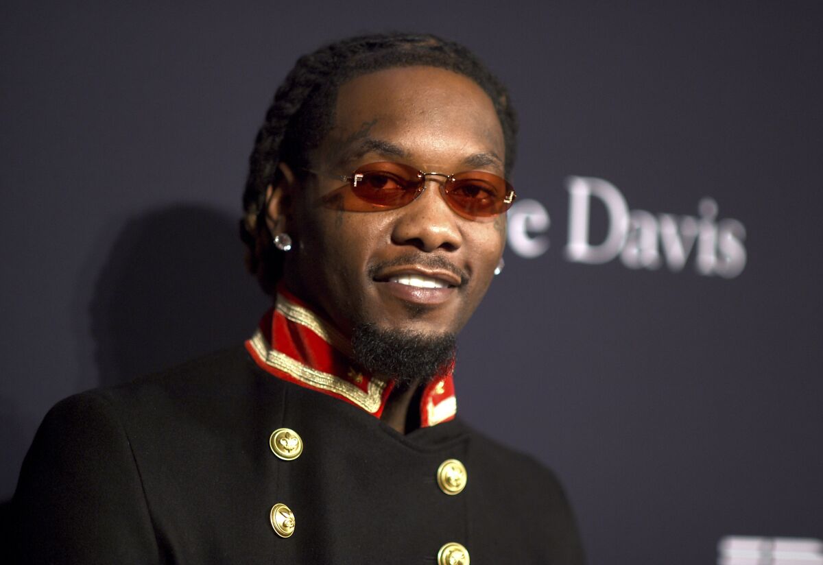 Offset smiles while wearing tinted red glasses, earrings and a dark jacket with gold buttons and a red collar.