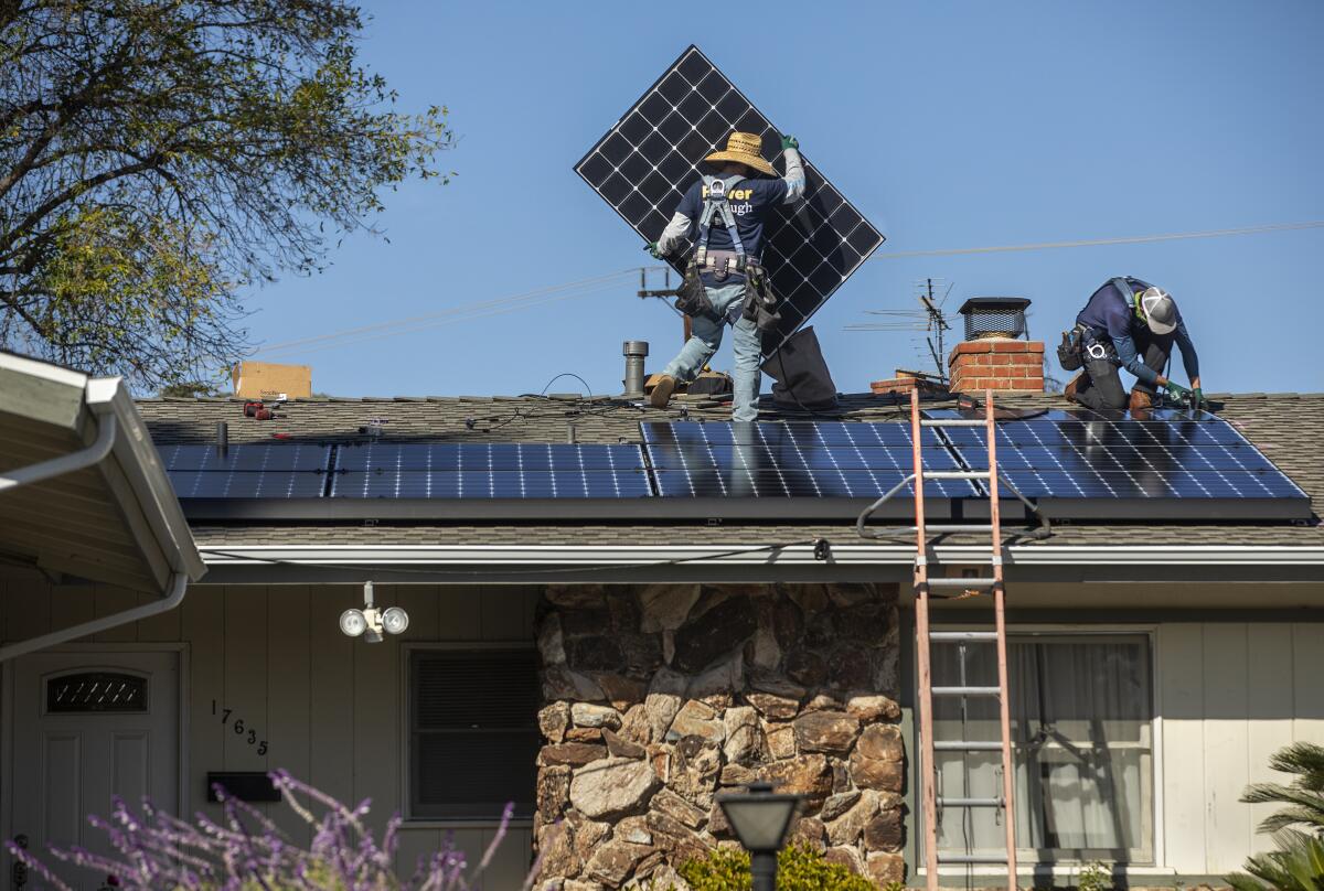 A ladder leans against a home where, atop the roof, two workers are carrying and placing solar panels.