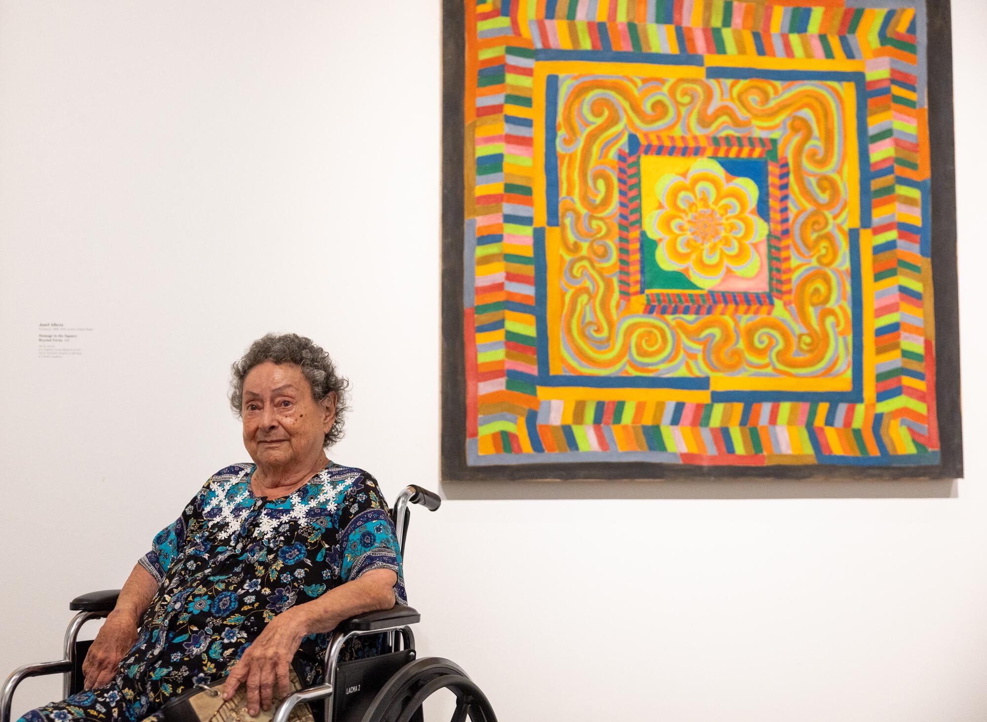 An older woman in a patterned top sits in wheelchair next to a painting that looks almost quilt-like in its pattern.