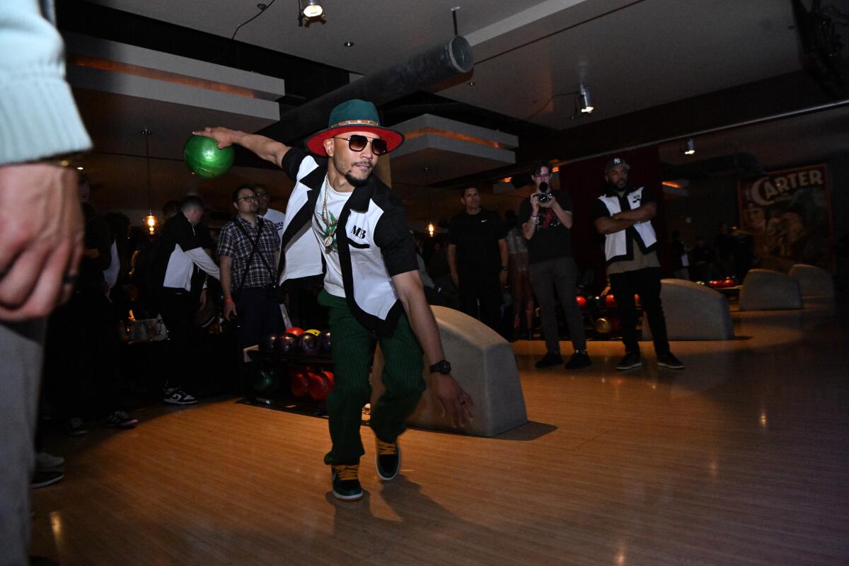 A man in shaded glasses with a green hat holds a green bowling ball in one hand behind him in a bowling alley.