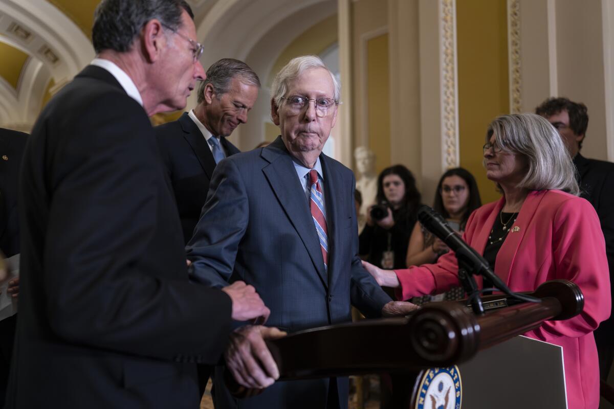 Colleagues take Mitch McConnell's arms as he stands at a lectern in an ornate room while others look on