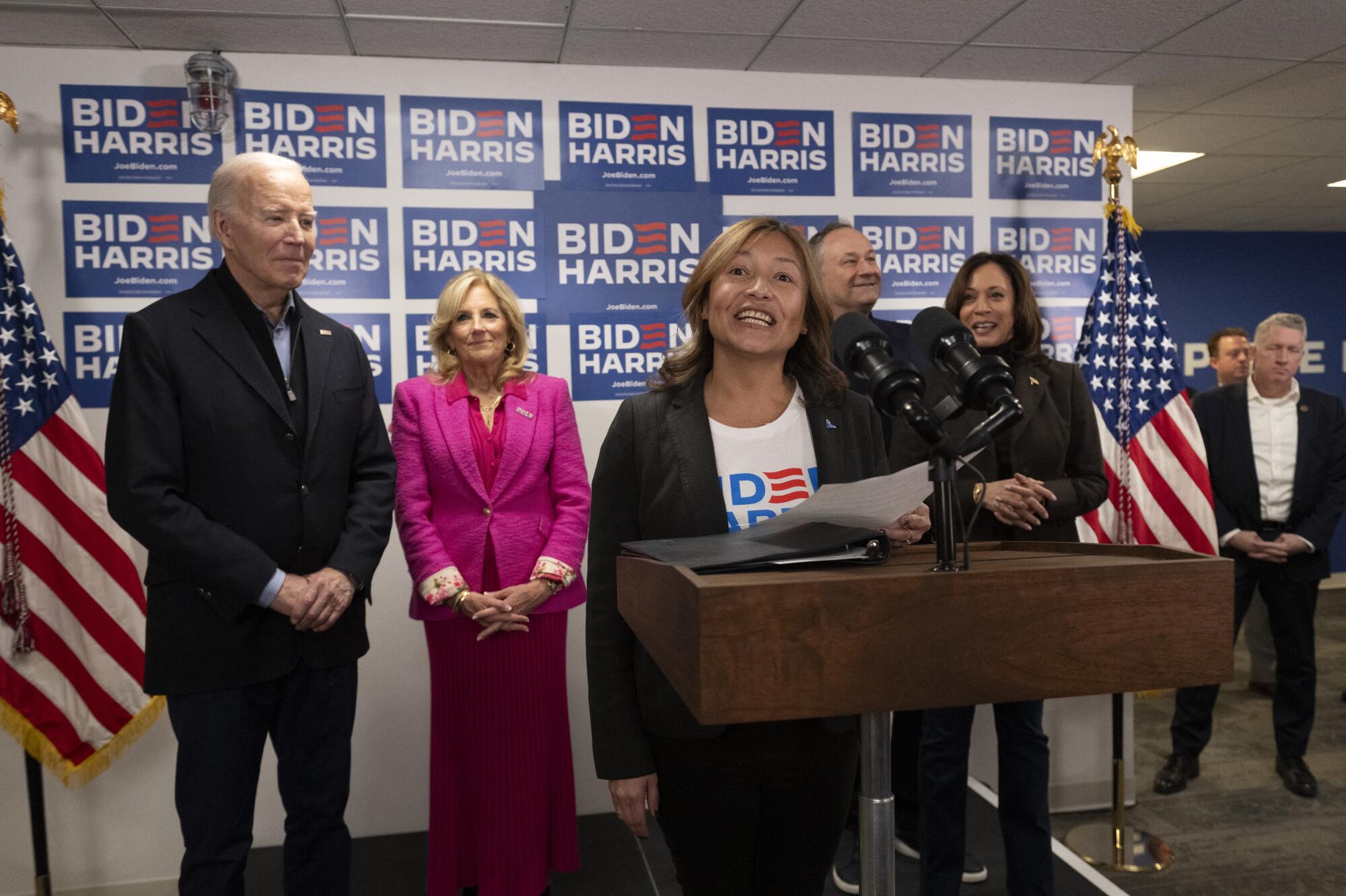 A woman smiles while speaking at a lectern, with two couples behind her, also smiling, and Biden Harris signs