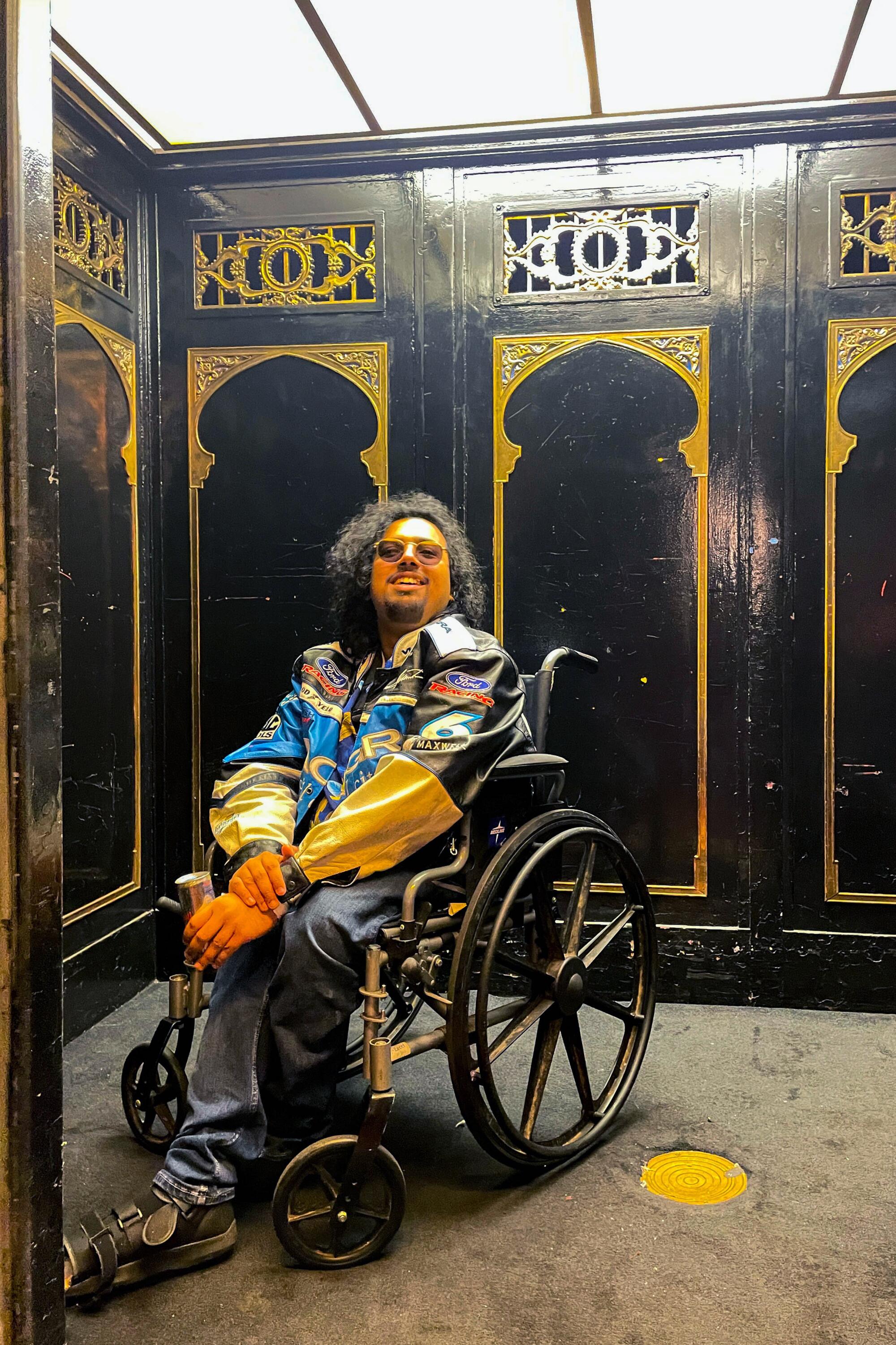 Saptarshi Majumdar wearing a racing jacket sits in a wheelchair in an elevator with black walls and gold trim. 