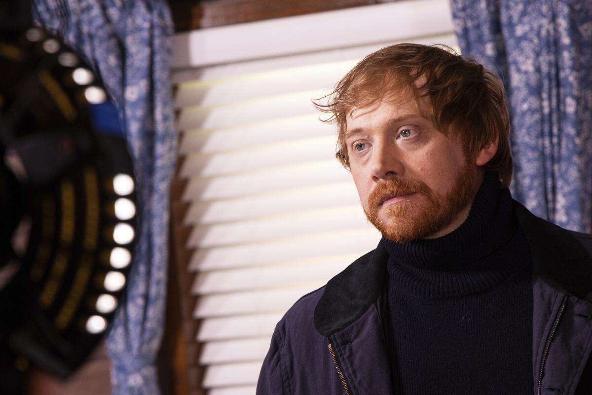 A redheaded man with a beard looks serious standing near a window with a blind and decorative curtains