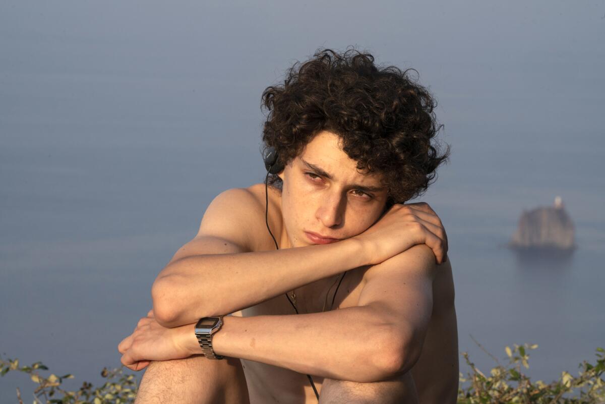 A young man sits by the water, looking contemplative, with his arms on his knees.