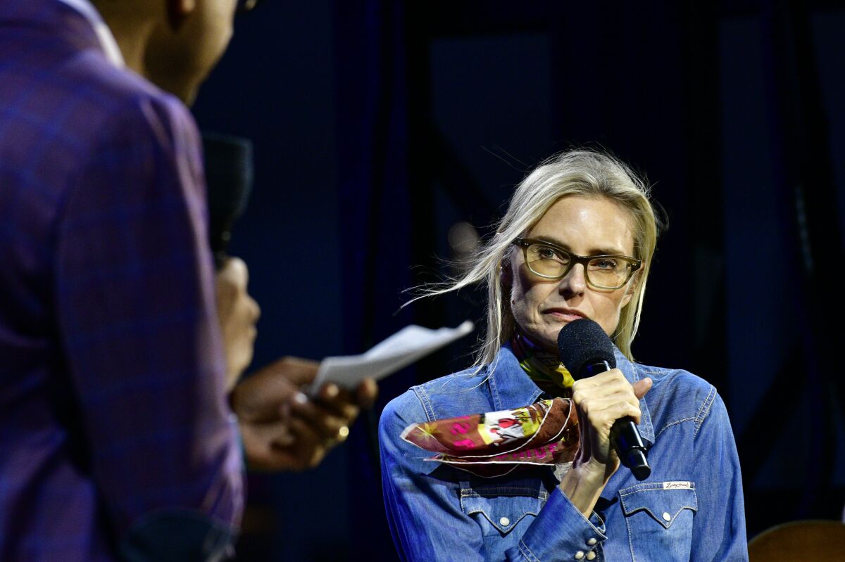 A woman in a denim shirt and glasses holds a microphone in front of her face