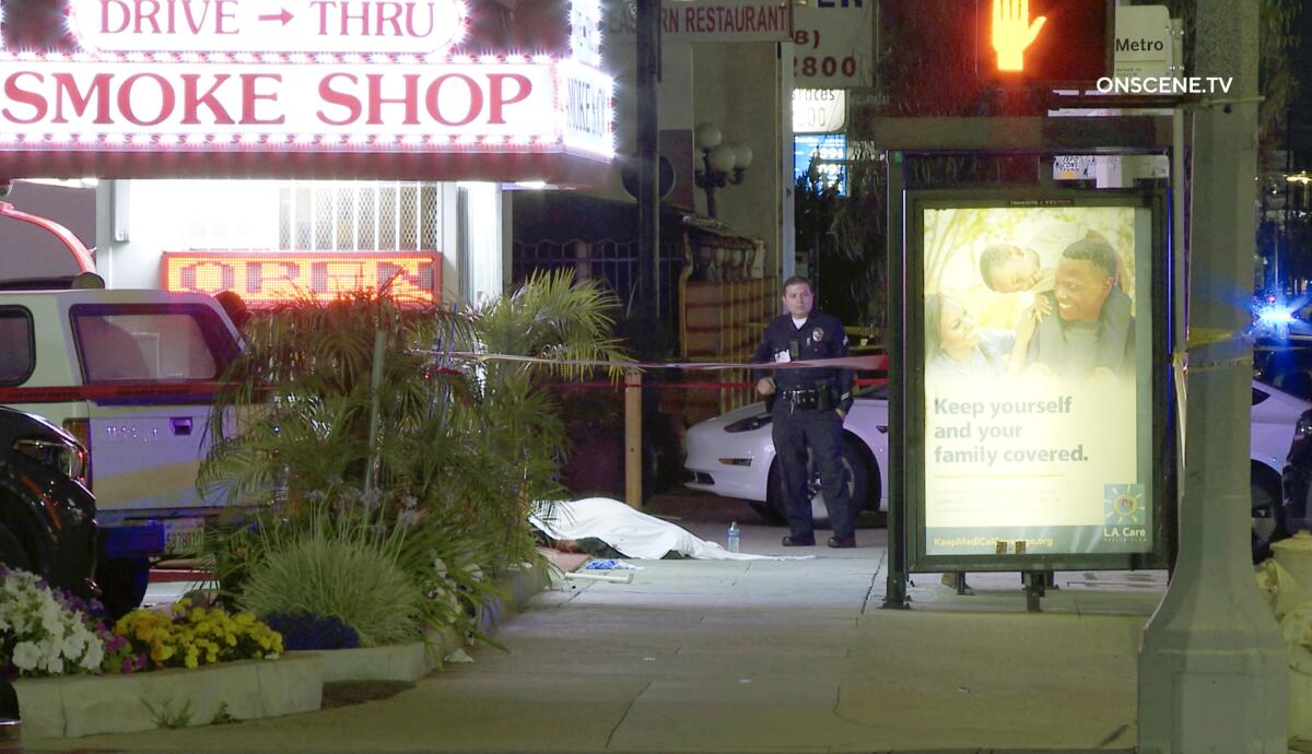 A police officer next to a crashed car and a body covered by a white sheet on a sidewalk outside a smoke shop