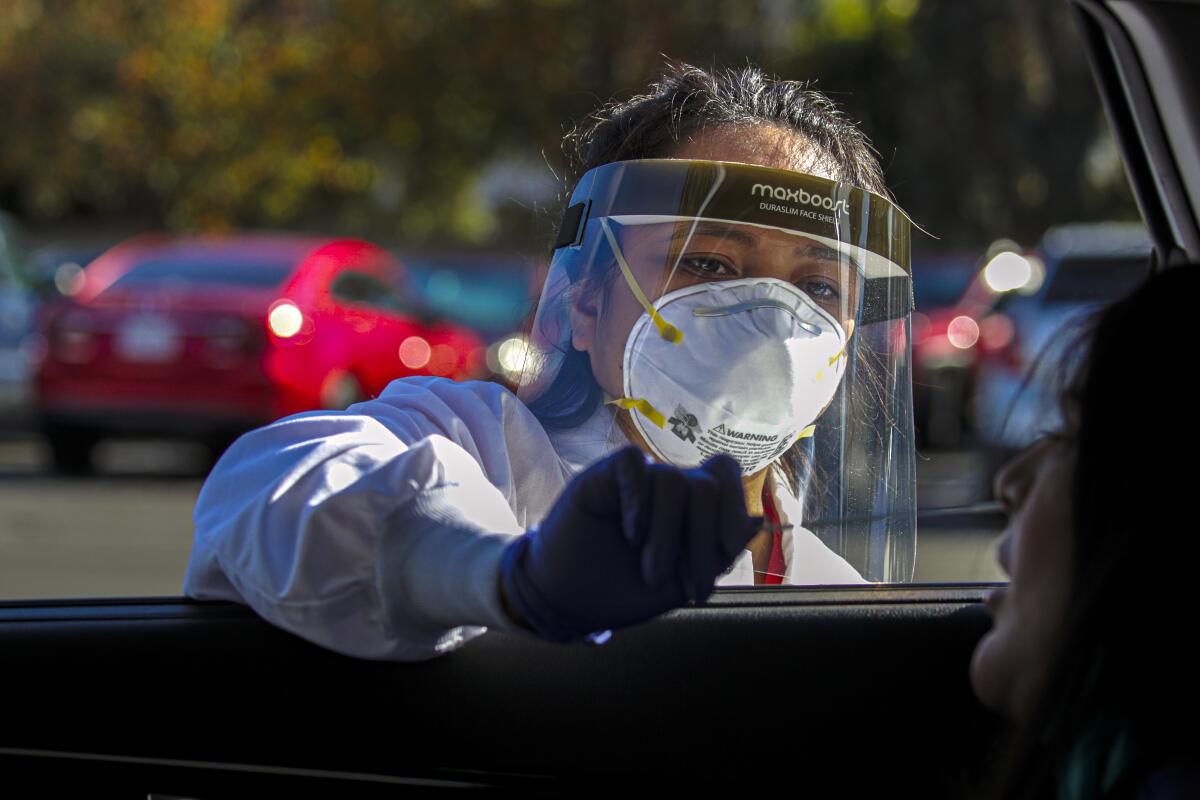 A nurse in face shield and mask reaches a gloved hand inside a car window to insert a long swab into the nose of a woman. 