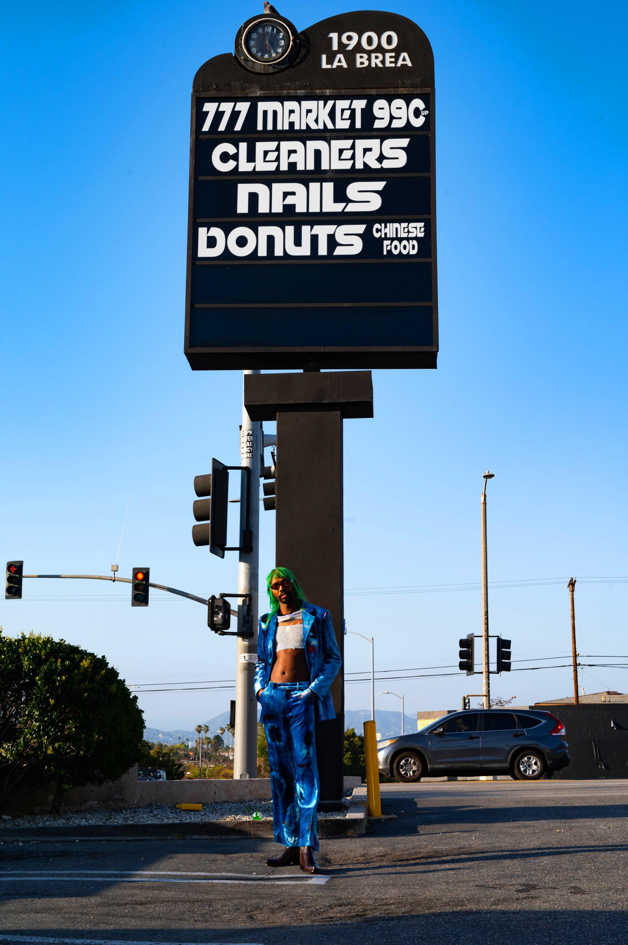 A person in a blue suit stands below a strip mall sign for 1900 La Brea.