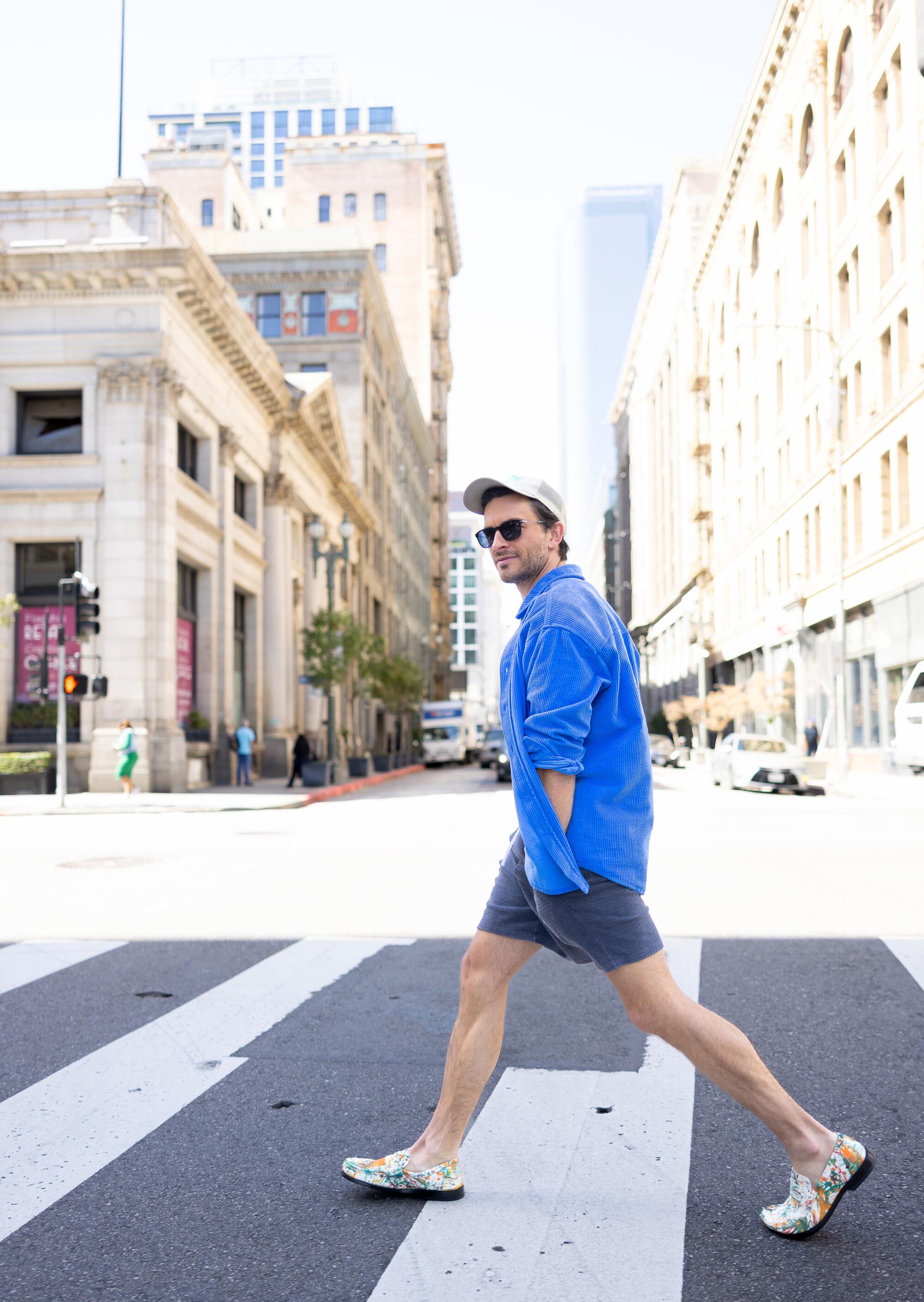 A man wearing a hat, blue T-shirt and shorts crosses a street in downtown Los Angeles.