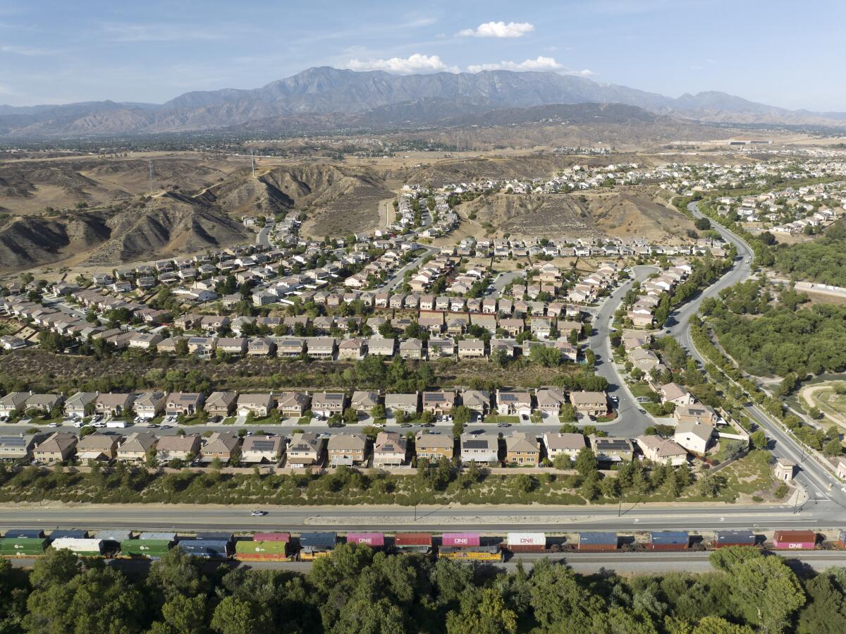 An aerial view of a housing development backed by mountains. A train passes along in the foreground.