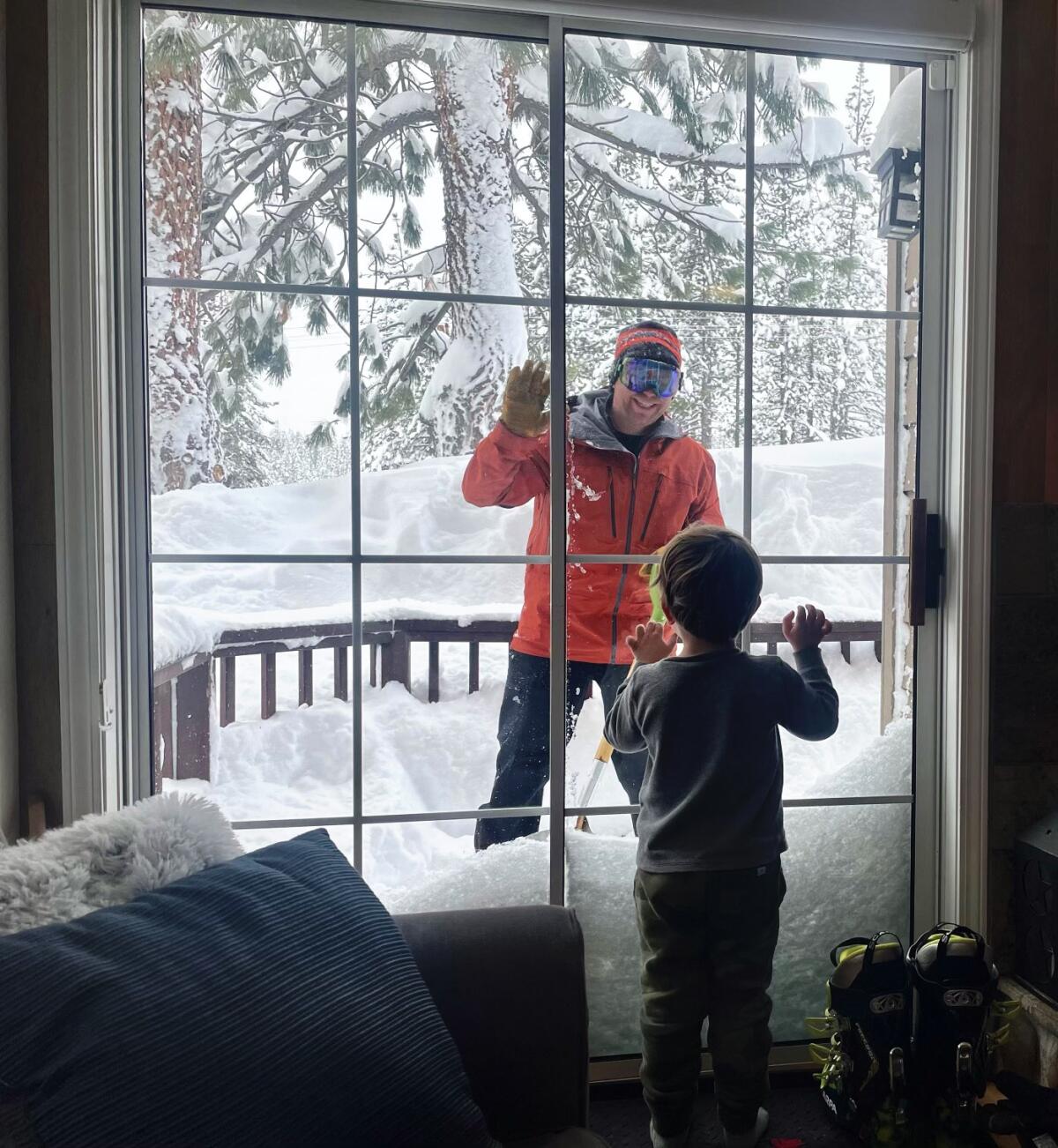 A man in ski gear waves from a snowy deck through a window to a child.