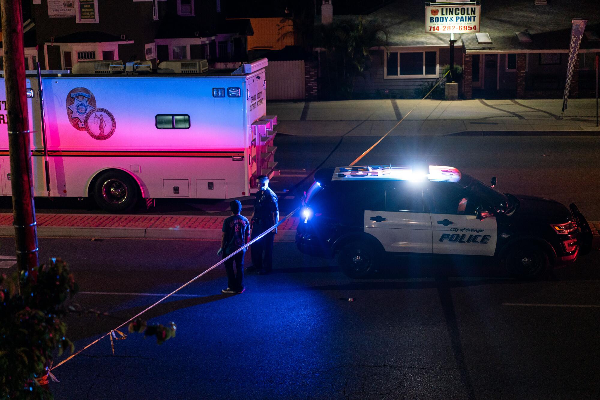 Two officers stand alongside crime tape in the glow of lights from a police car.