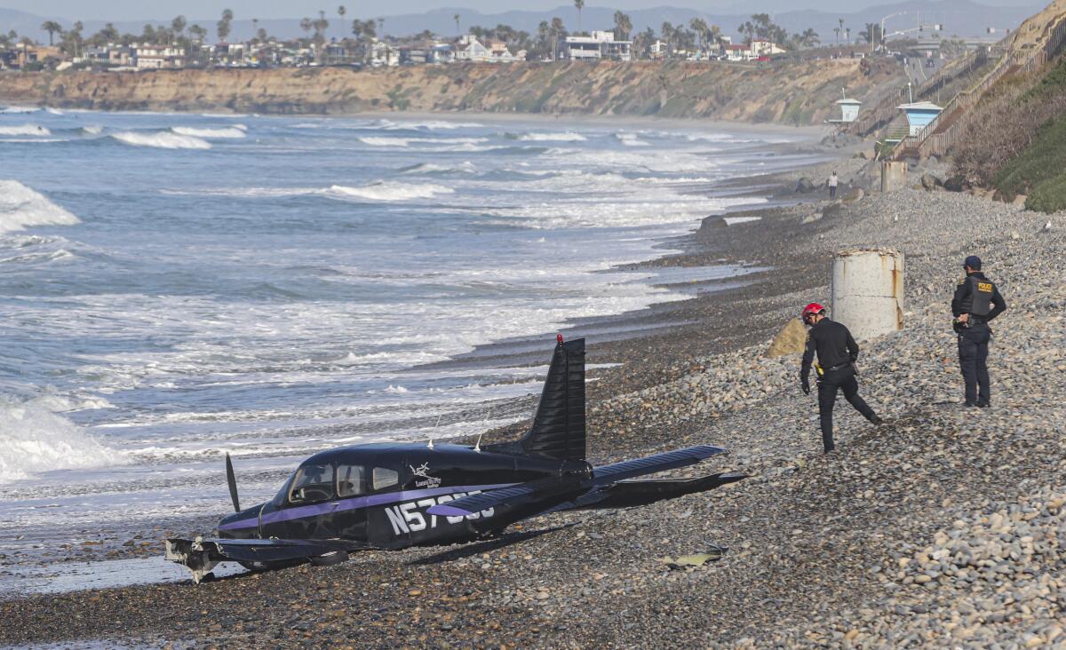 A small single-engine plane with a damaged left wing sits on a beach in the surf line