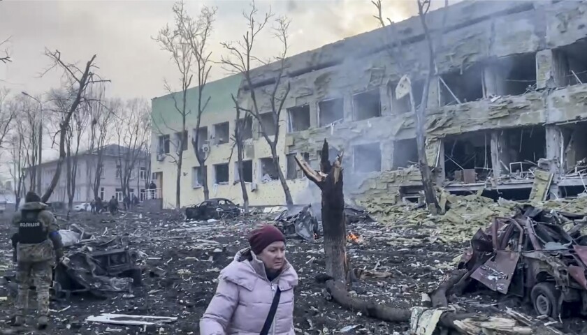 A person walks past bombed buildings and burned trees.