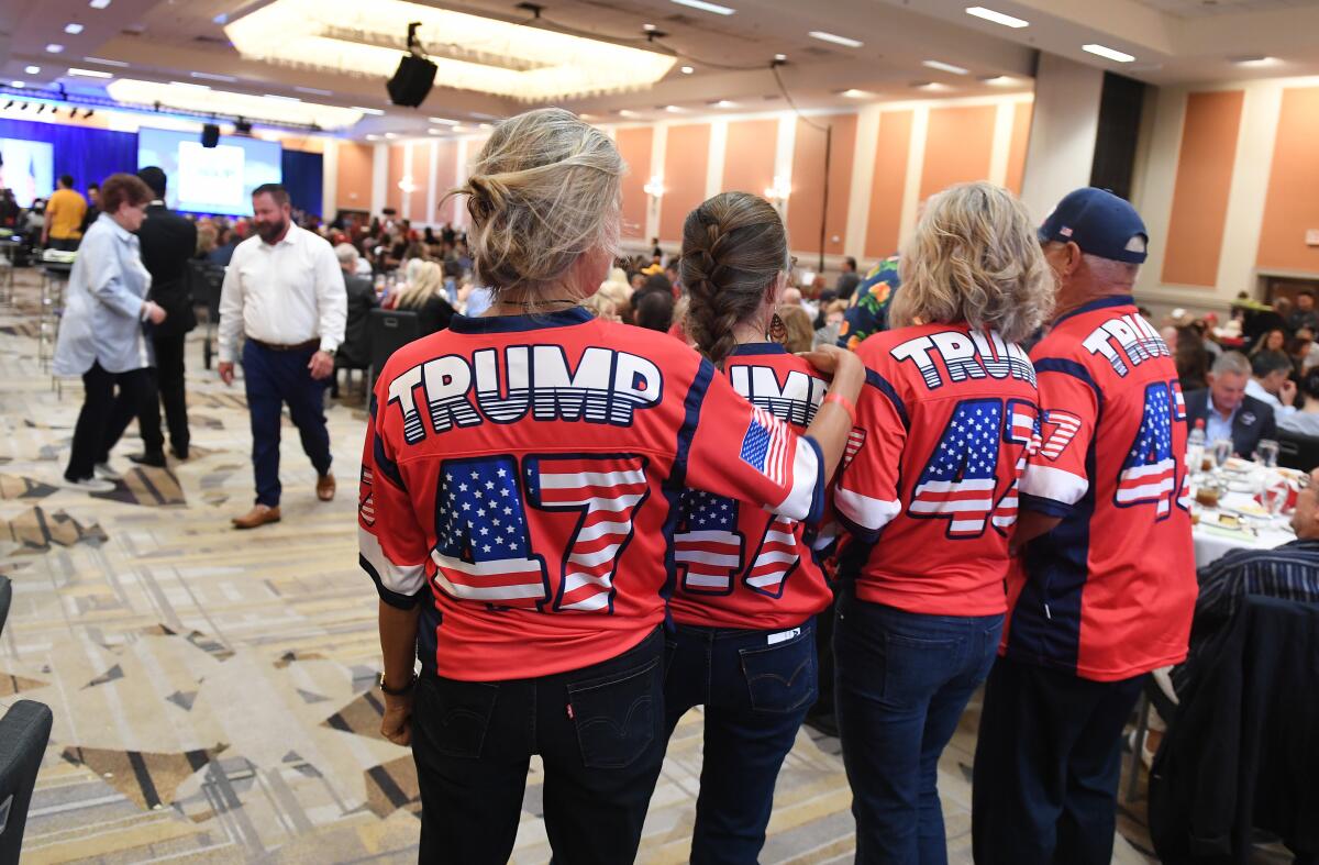 Four people, seen from behind, wearing red shirts with the words Trump 47, stand in a room with people seated at tables 