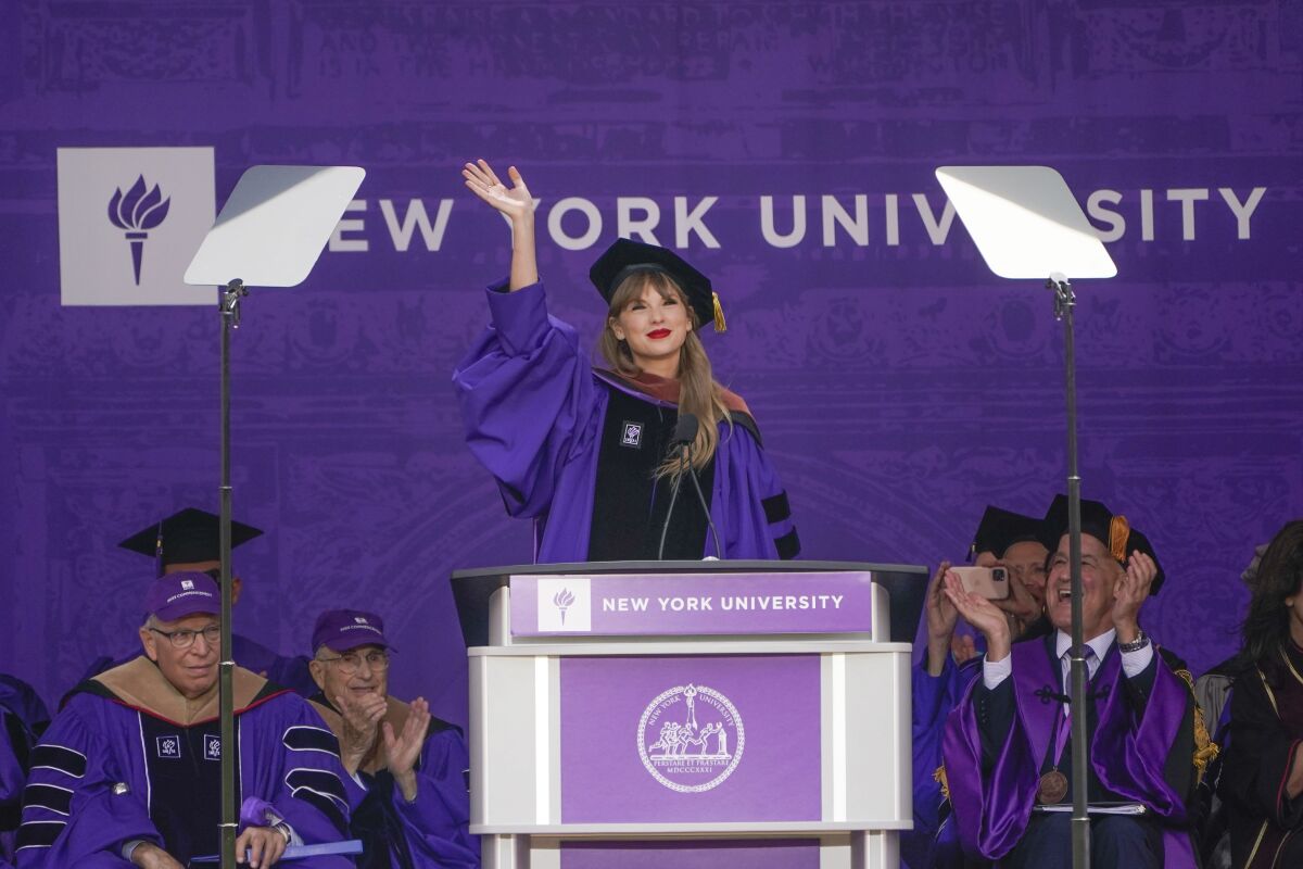 A blond woman in a graduation cap and gown waving from behind a podium on a stage