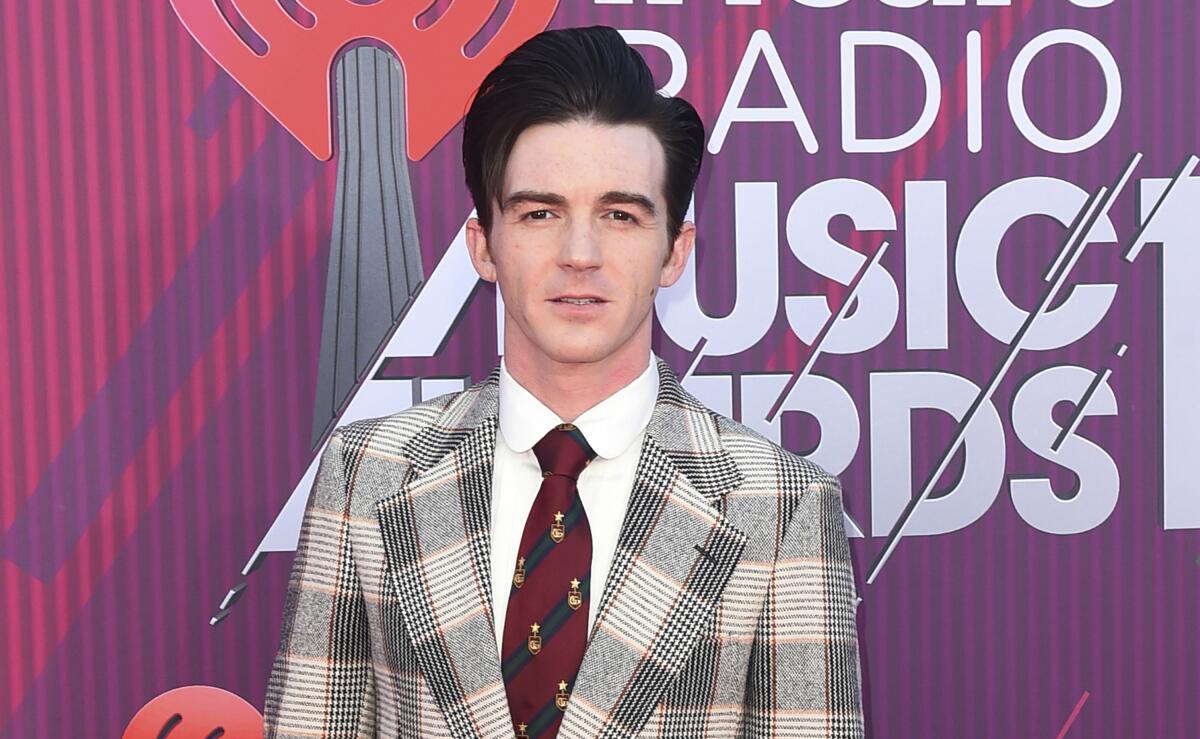 A man in a gray and beige plaid suit with a red tie stands against a purple and red backdrop with white text