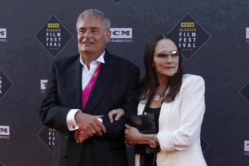 Leonard Whiting, in a black suit, links arms with Olivia Hussey, in a white suit, as the two pose with slight smiles.