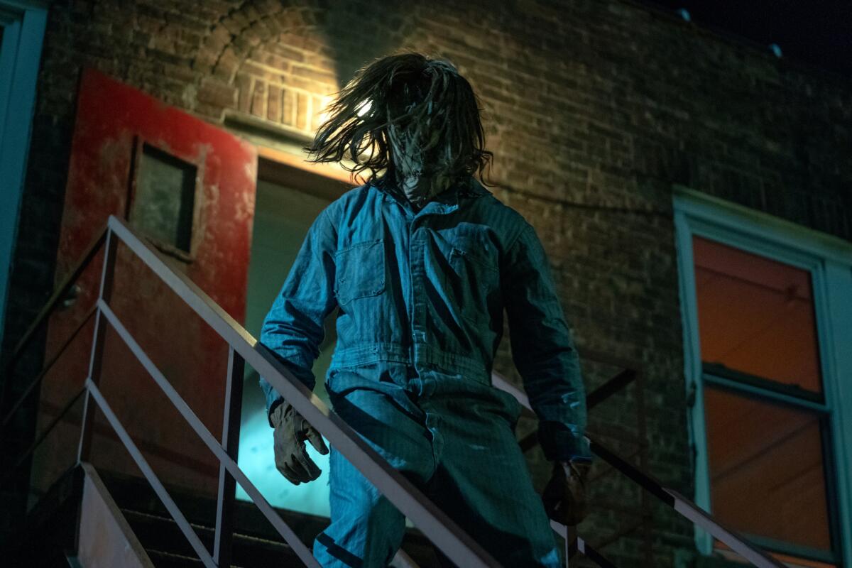 A killer in a mask, gloves, long hair and jumpsuit standing on a fire escape.