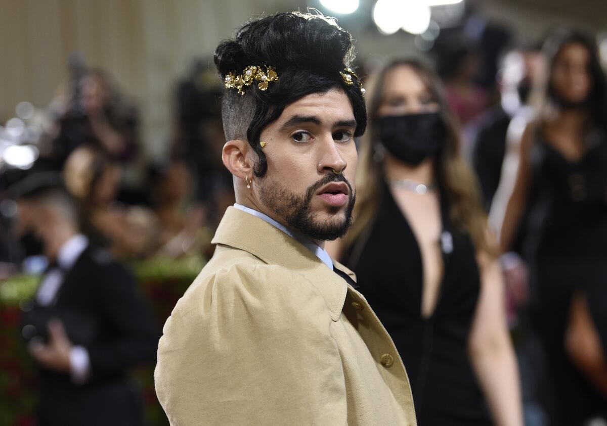 A man wearing his black hair in a bun atop his head poses in a beige coat