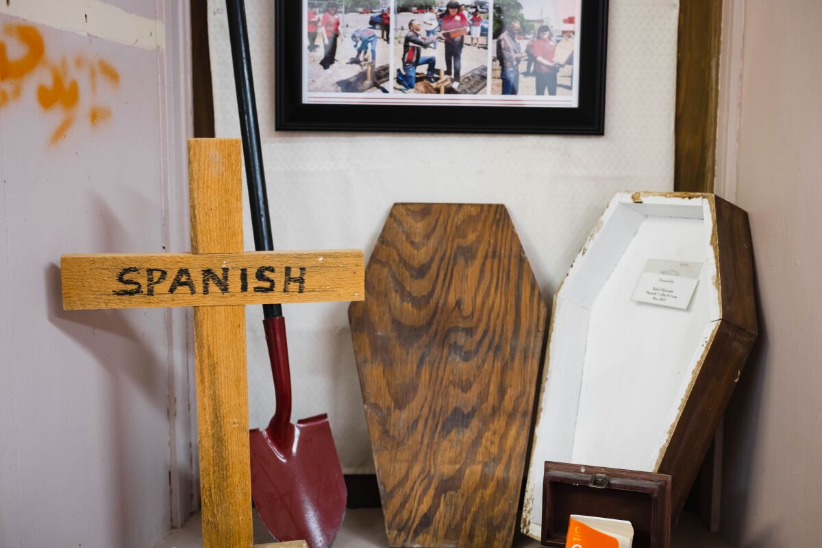 A wooden cross bearing the word "Spanish," a shovel and a small open coffin on display near a framed collage of photos 