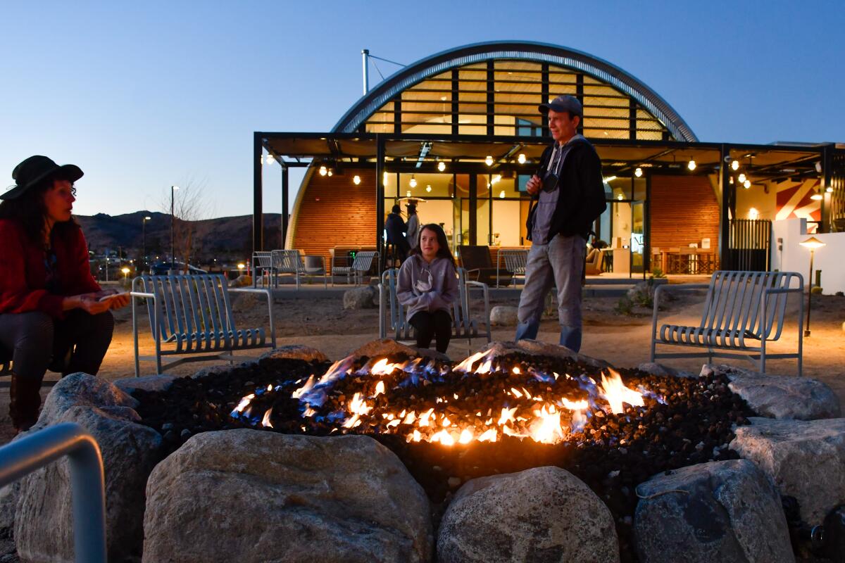 Three people sit around a large campfire at dusk. In the back is a modern-looking building with rounded exterior.