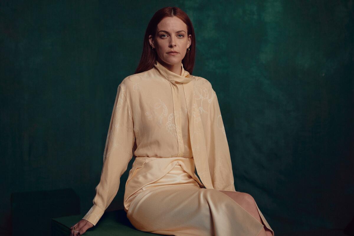 Riley Keough seated with arms to her sides in a peach long-sleeve blouse and skirt
