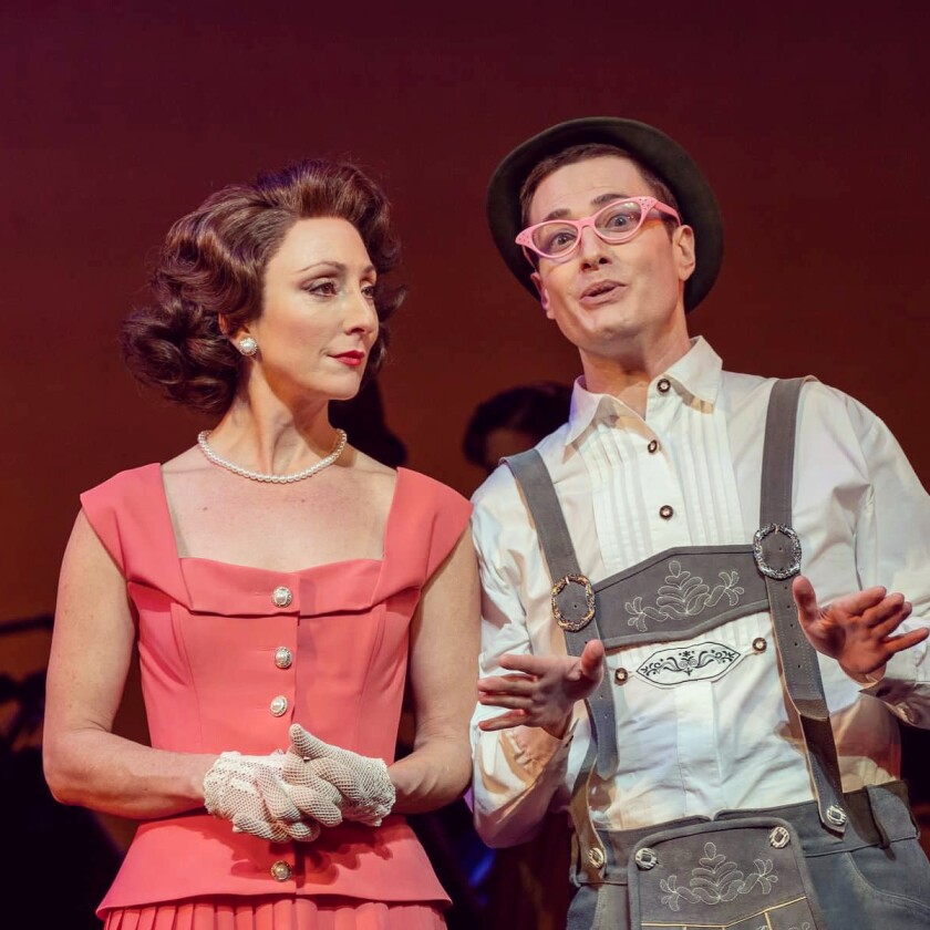 A woman in a salmon-colored dress and a man in pin kglasses and Lederhosen in “Call Me Madam.”