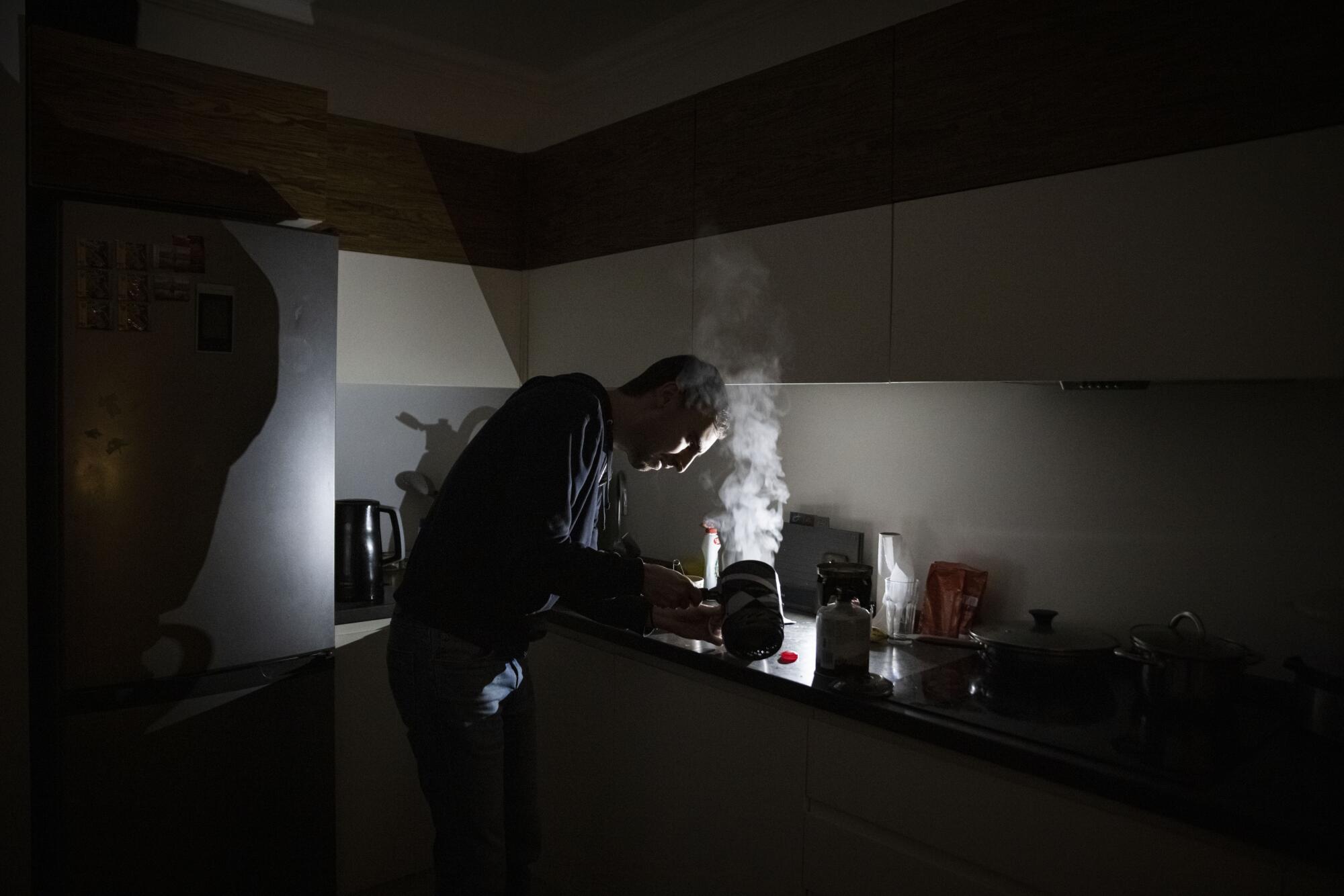 A man makes tea on a camping stove in a dark kitchen.