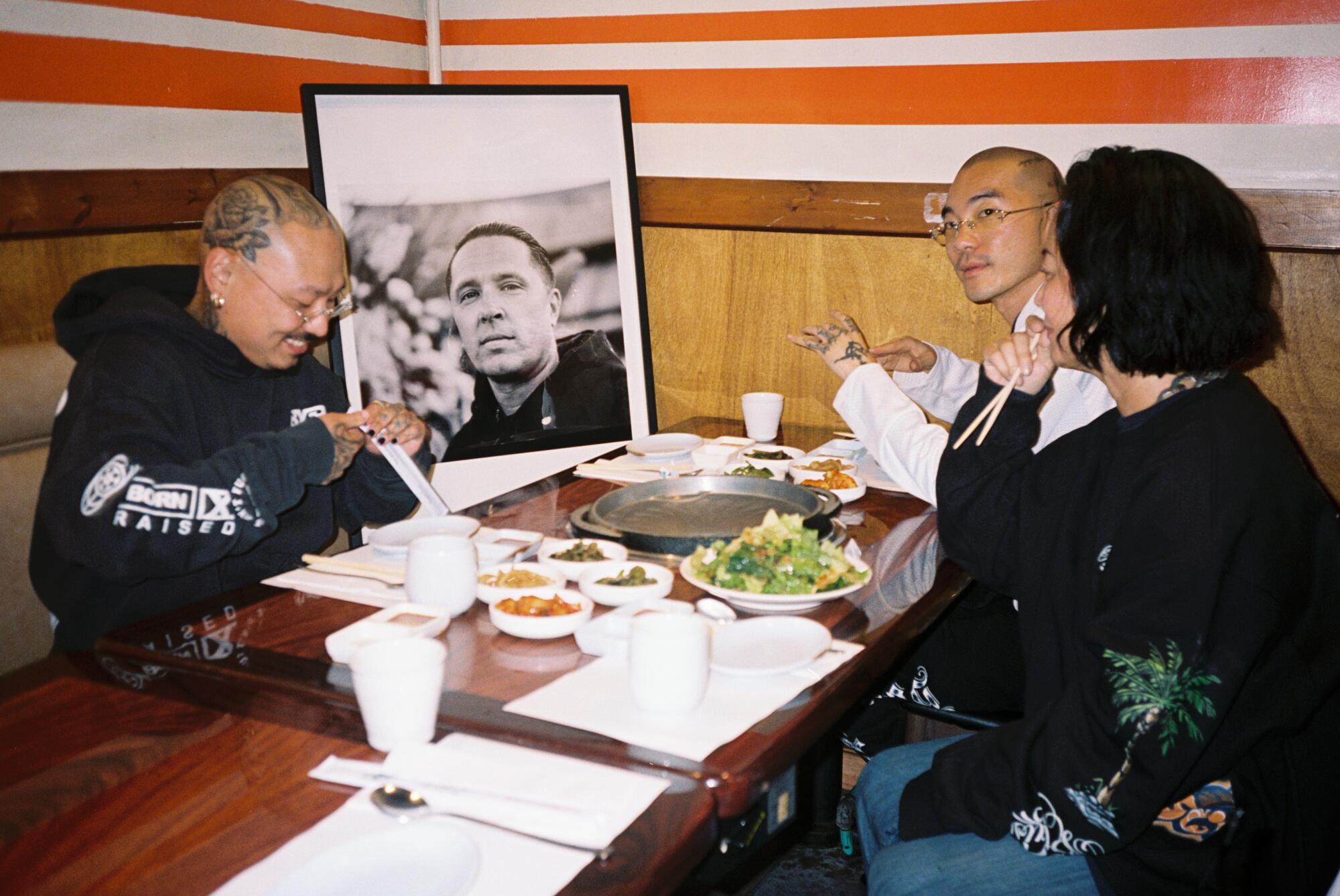 Three people sit and eat at a restaurant, with a black-and-white photo of a man propped up at the table.