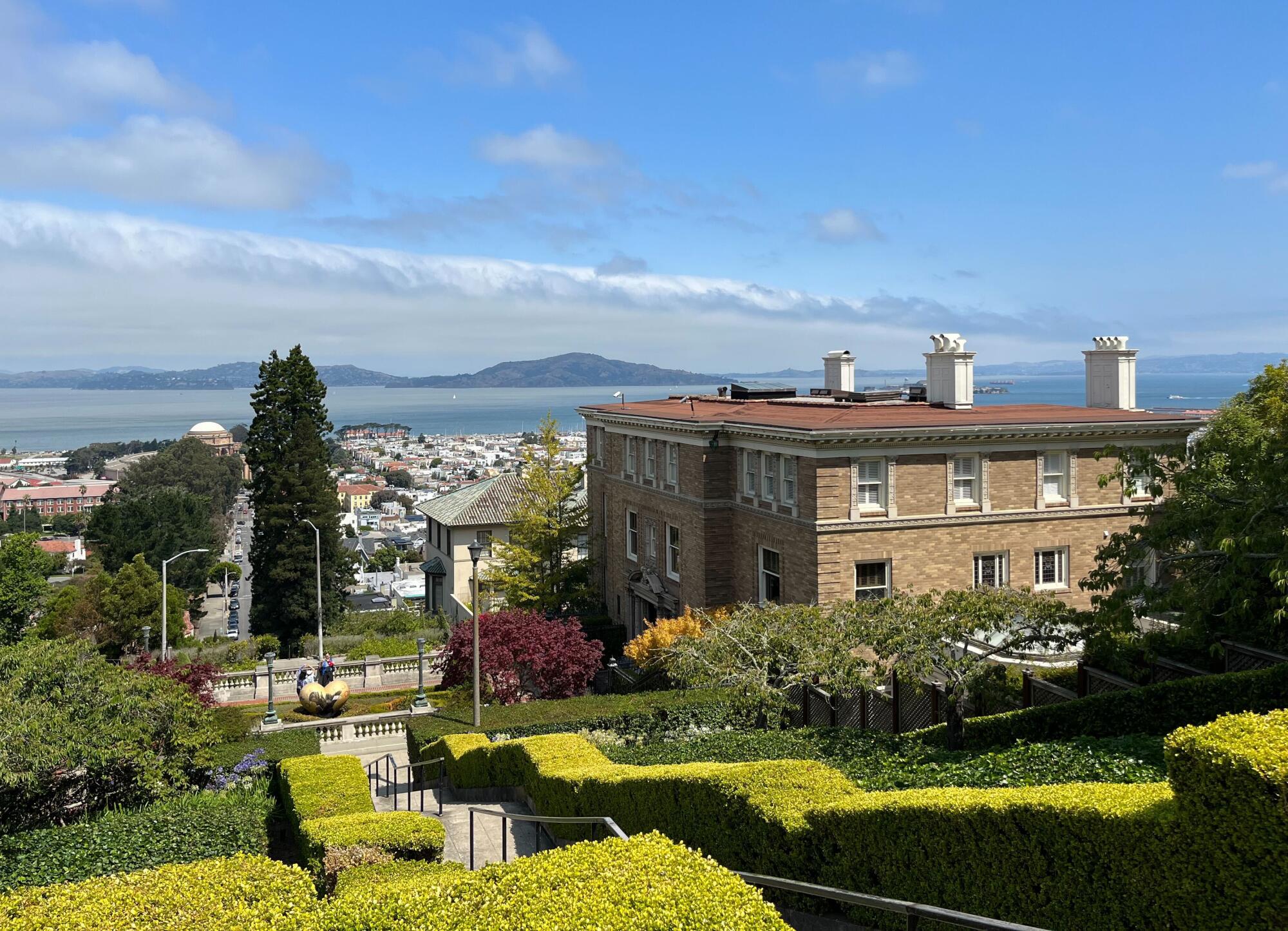 Trimmed shrubs line a hillside staircase next to a brick mansion that overlooks city, water and mountains