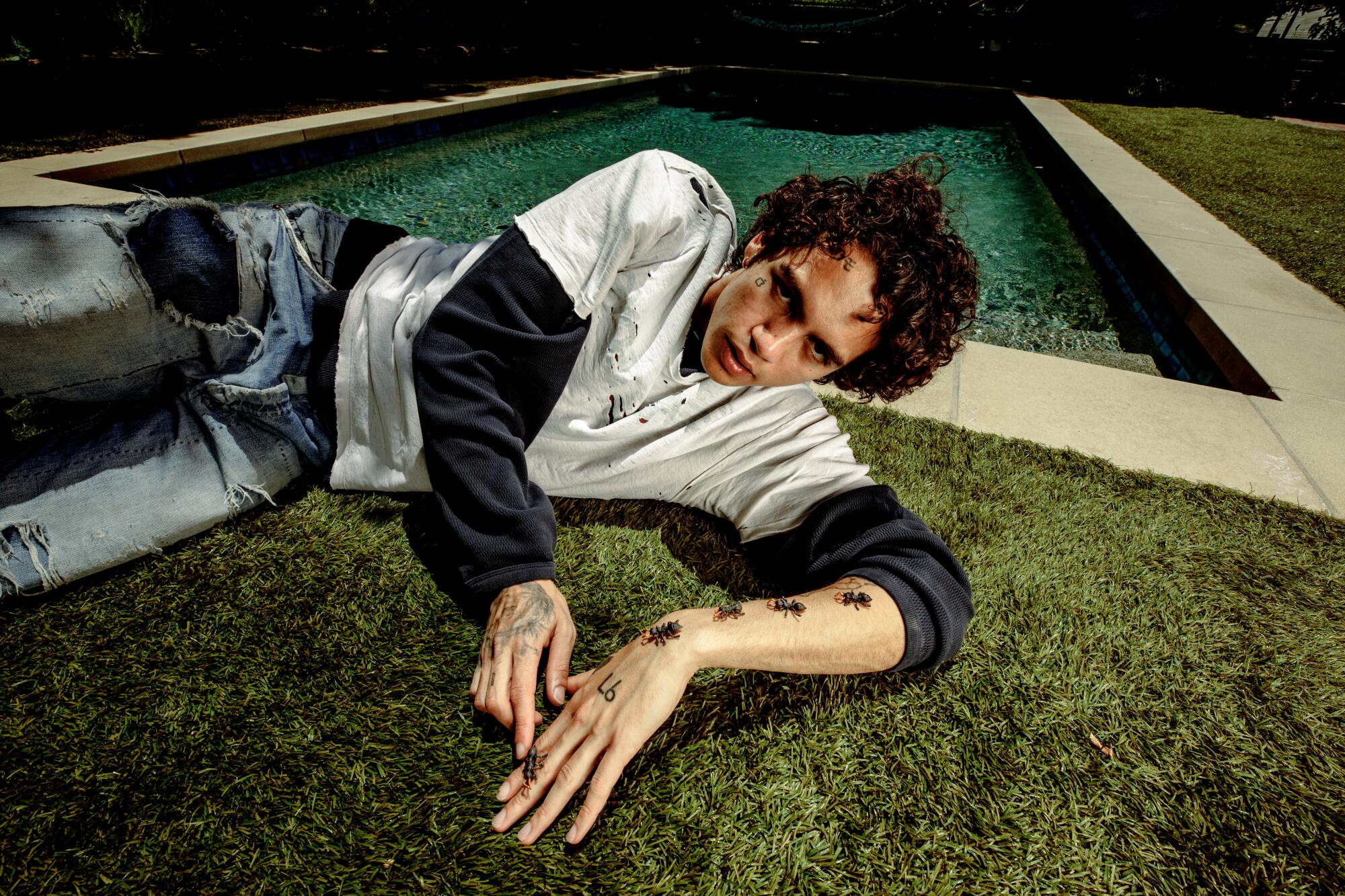 Dominic Fike lies on grass with fake ants crawling up his arm. 