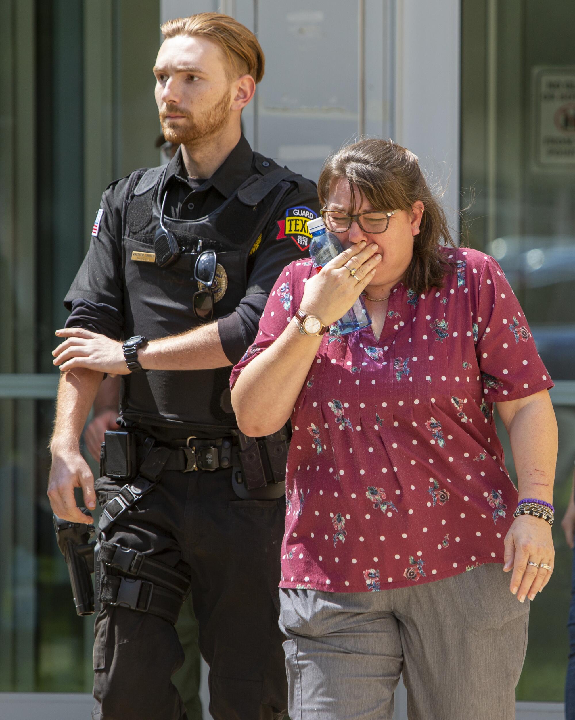 A woman in glasses holds a hand to her face as she cries while standing next to an officer in dark uniform 