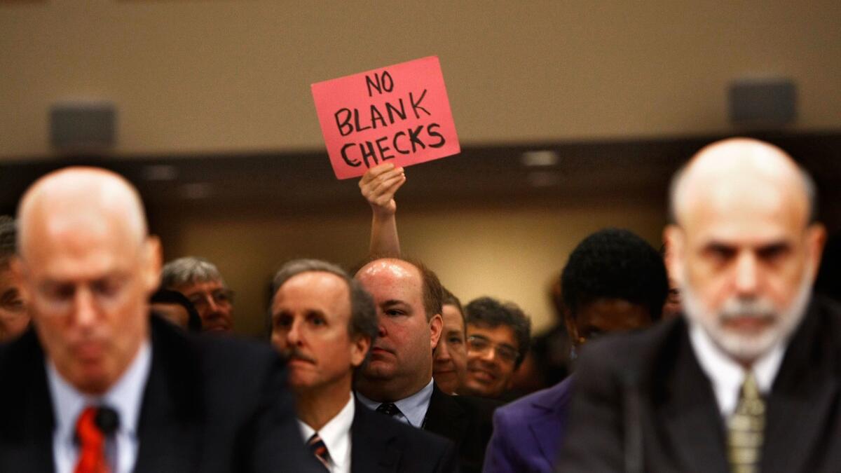 A demonstrator holds up a sign behind Treasury Secretary Henry Paulson, left, and Federal Reserve Chairman Ben S. Bernanke, right, during a congressional hearing about the bank bailout fund in September 2008.