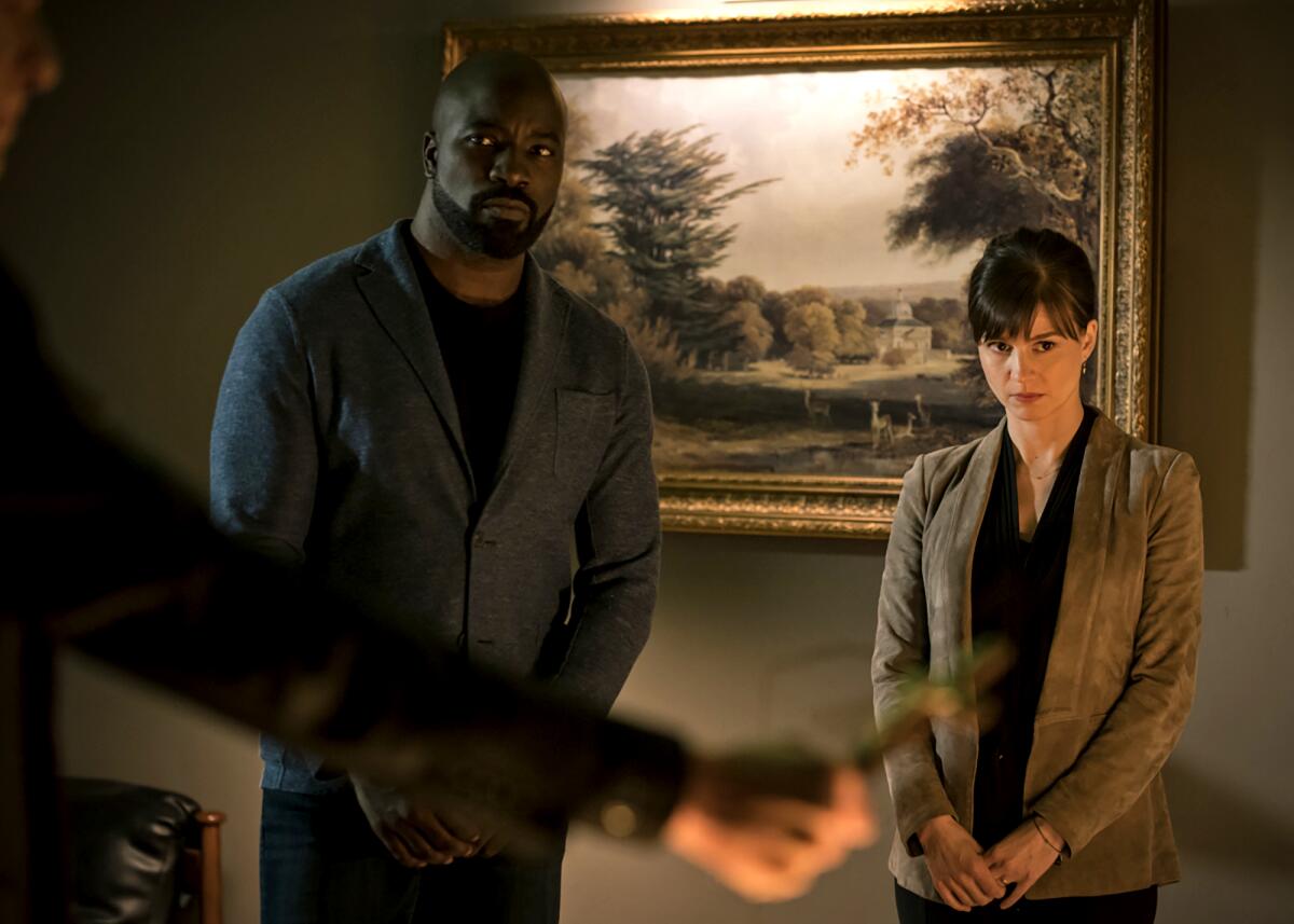 Mike Colter and Katja Herbers stand in a dim room in front of a landscape painting in the thriller "Evil."