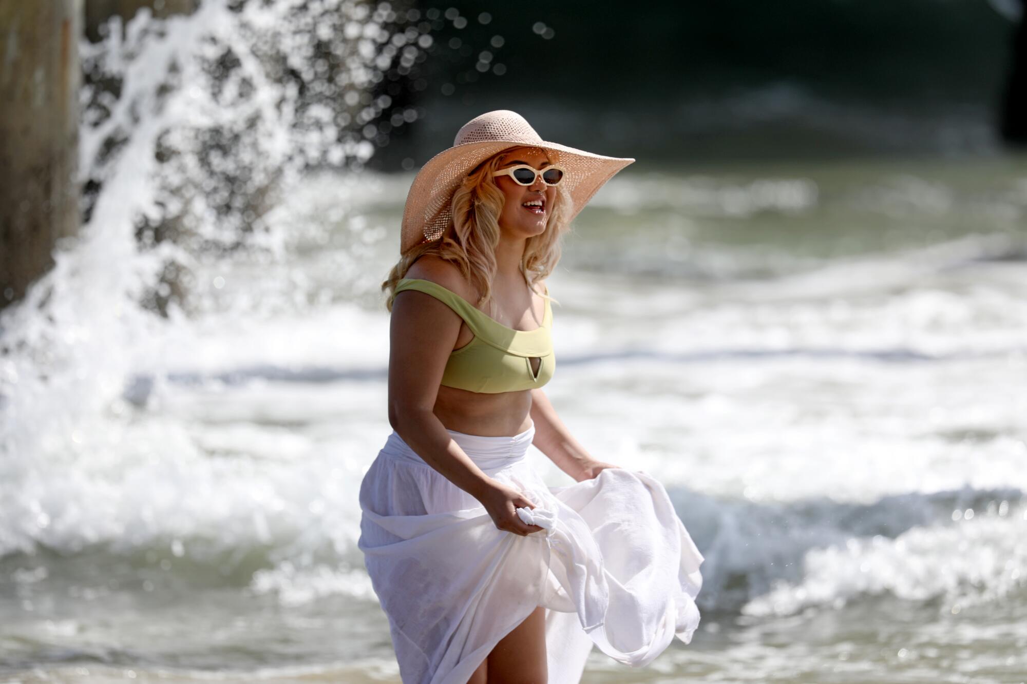 A woman in a hat wearing a green cropped top and white skirt stands with surf in background