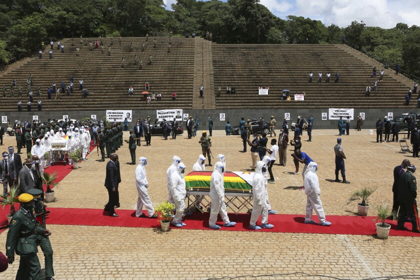 Amid military and other officials in masks, pallbearers in full hazmat suits carry coffins along a red carpet.