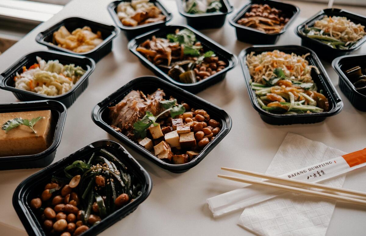 A photo of a range of Taiwanese dishes in balck to-go containers on a white table.