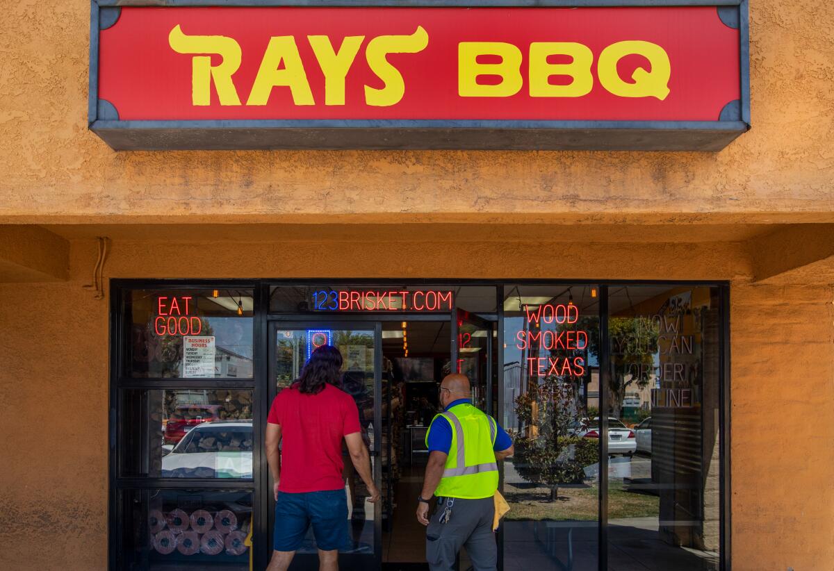 Two men walk into an orange stucco building under a sign that says "Ray's BBQ"